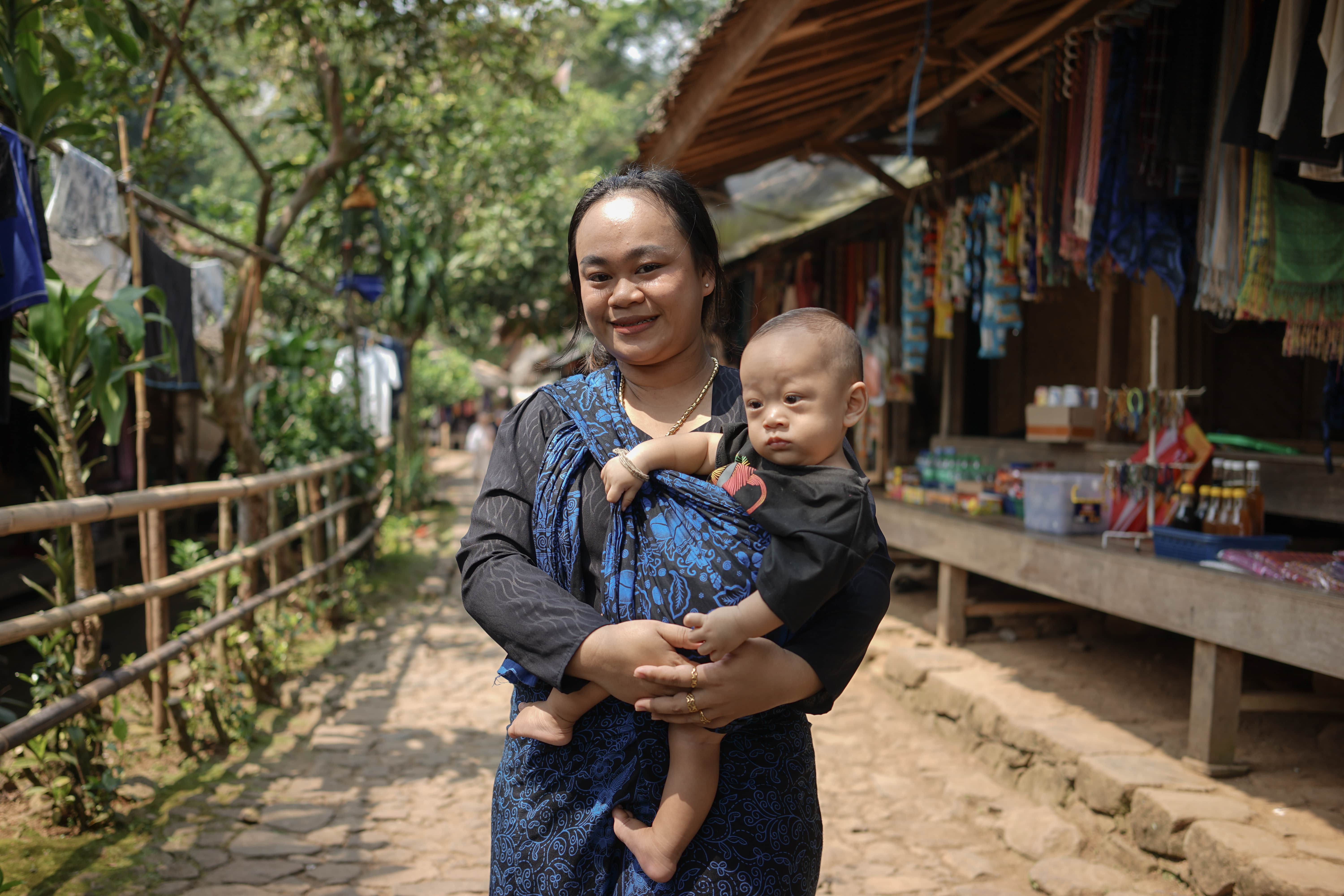 A woman wearing a black and blue kebaya holding a baby in front of a traditional shop. The background shows a village path with wooden houses, trees, and a peaceful rural atmosphere.